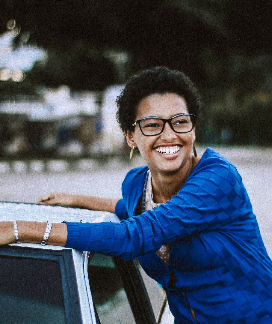 Young woman with short curly hair and glasses in a blue sweater standing next to her car.