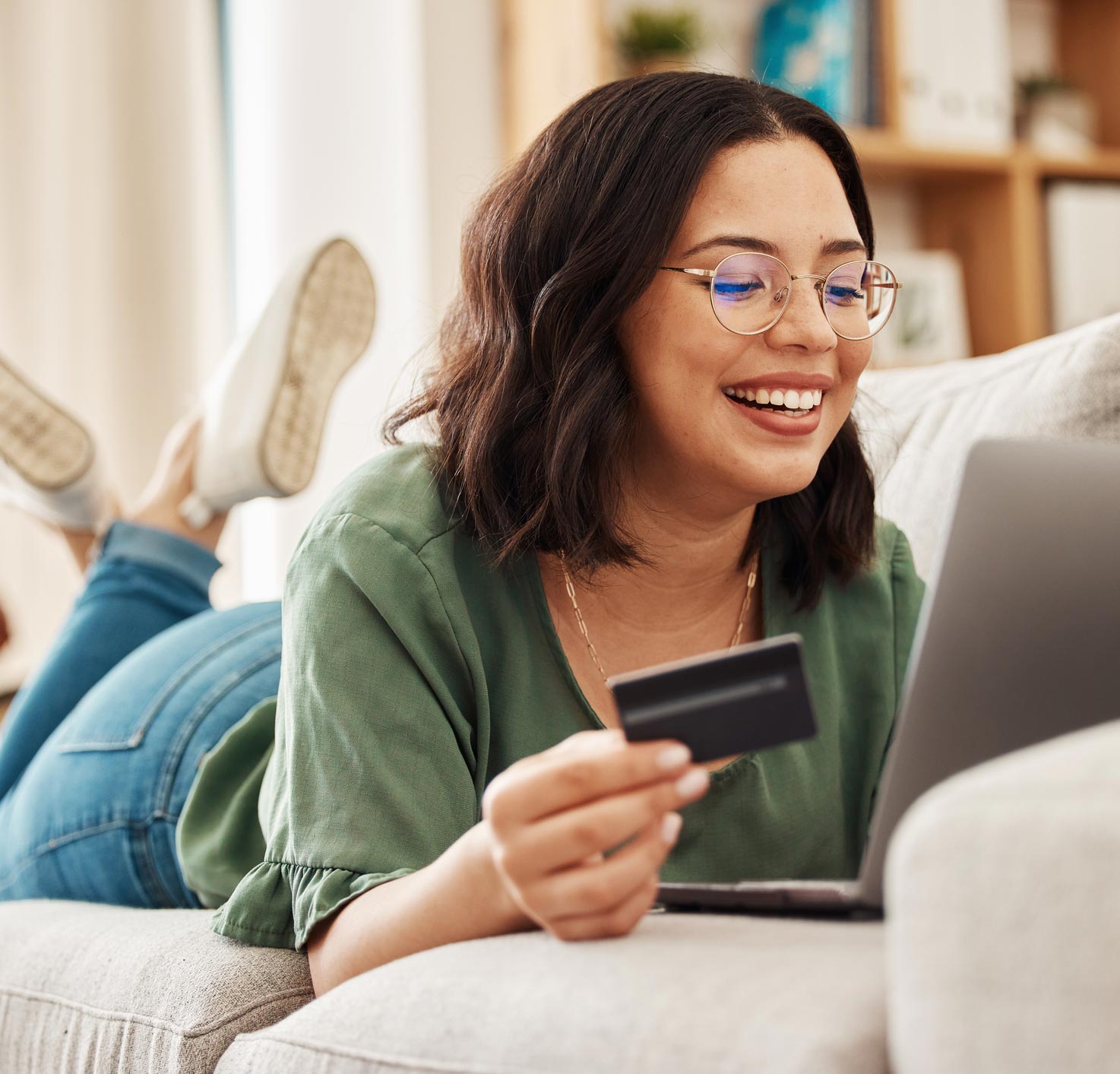 A woman wearing glasses holding a credit card and looking at her laptop on the couch.