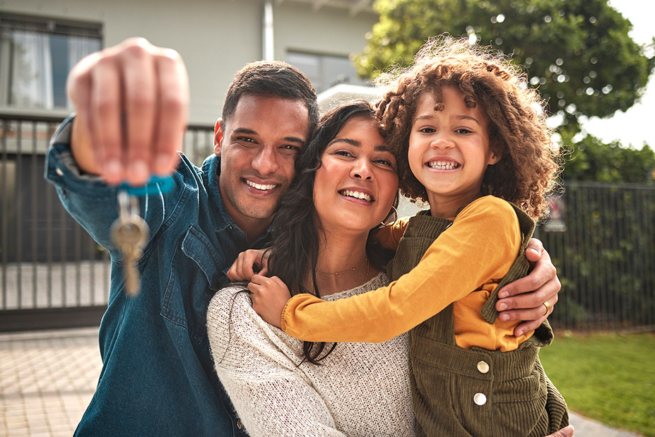 Happy family of three in front of their new house while Dad holds the keys and everyone including Mom and their child are smiling.