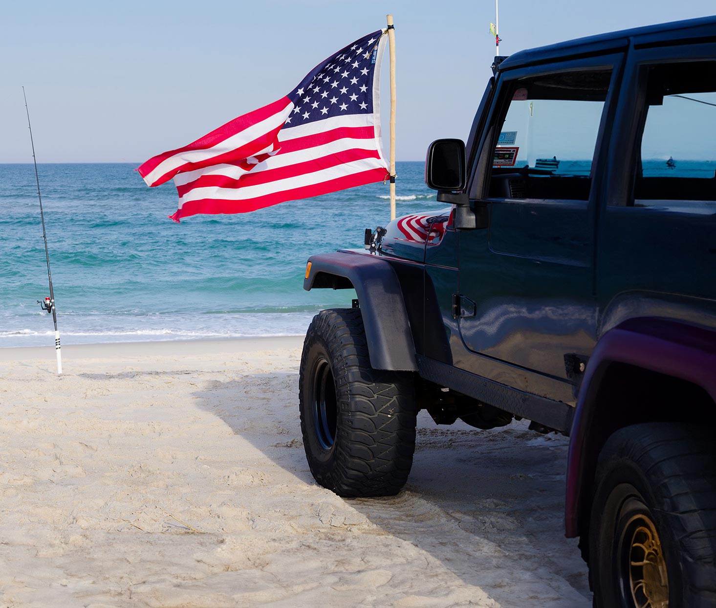 Blue jeep parked on the beach with an American flag and a fishing pole in the sand.