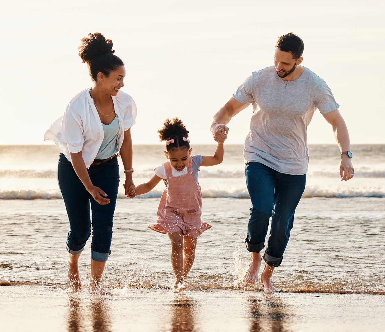 A mother, father and young daughter play joyfully on the beach at dusk with their clothes on but no shoes.