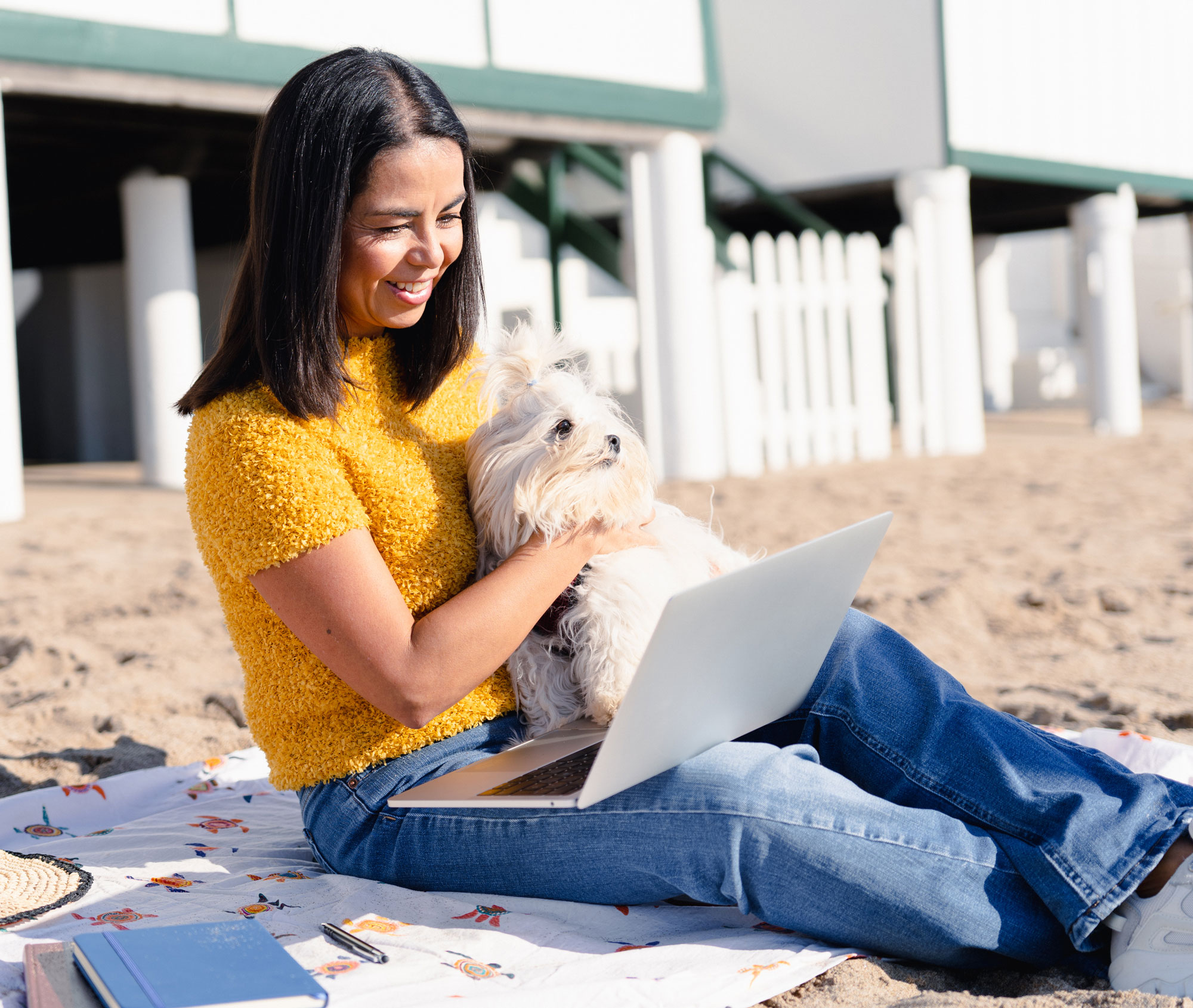 A smiling woman on the beach holding her dog and looking at her laptop viewing her money market account online.