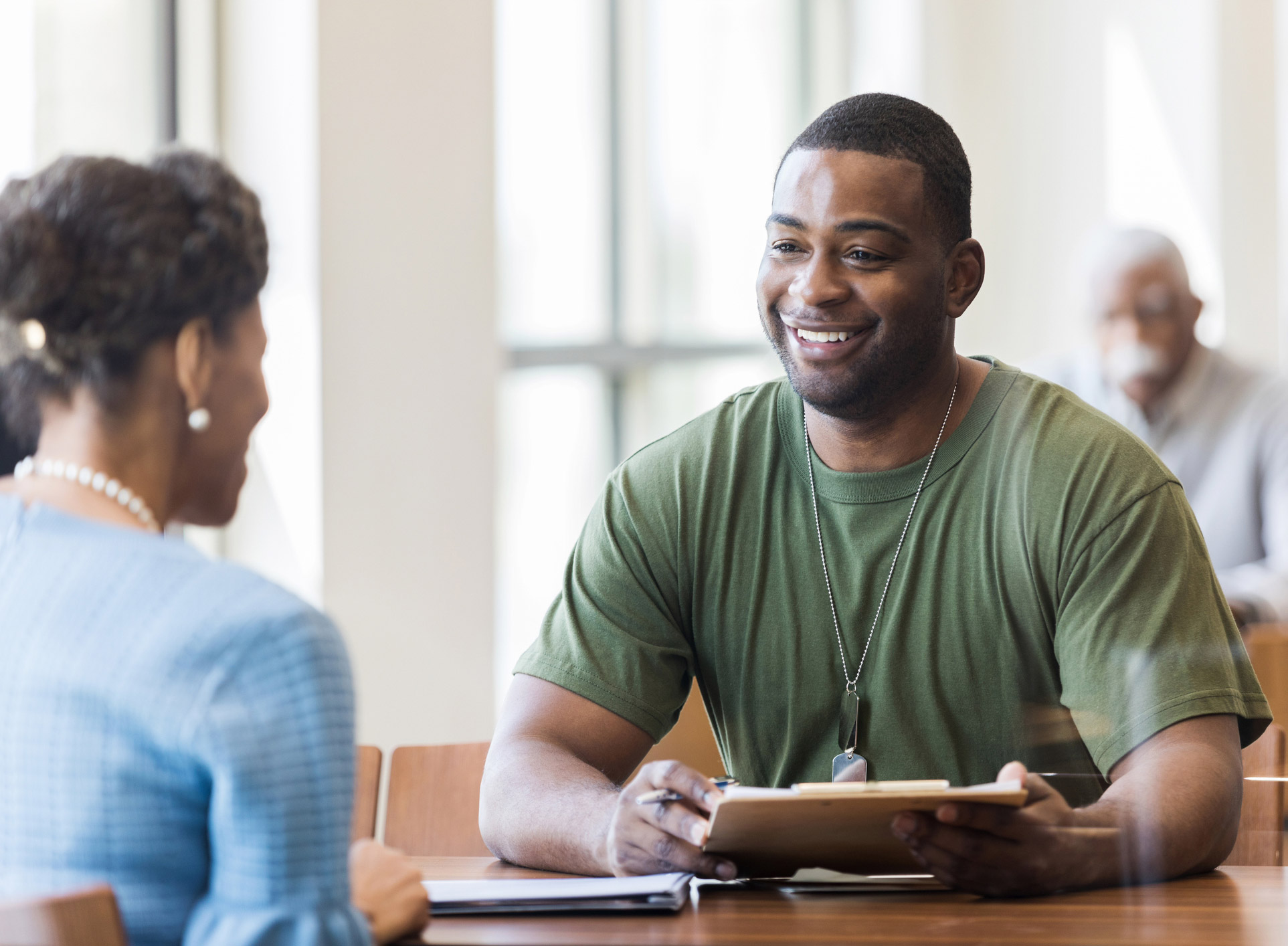 Woman in business attire talking to a male veteran wearing a military green t-shirt about his financial goals.