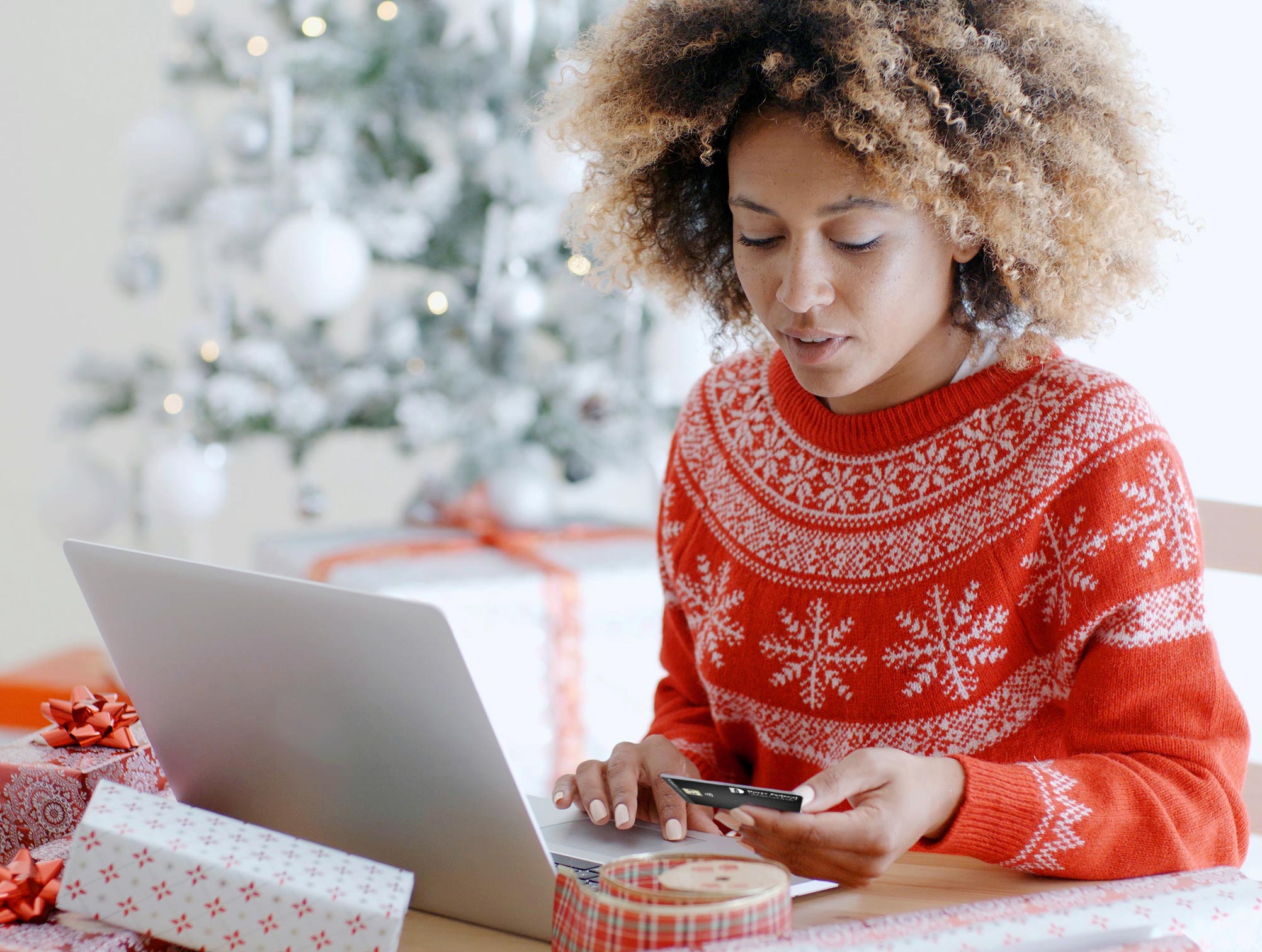 Woman in a holiday sweater in front of her laptop surrounded by presents looking at her Dover Federal Credit Union Cash Back Mastercard.