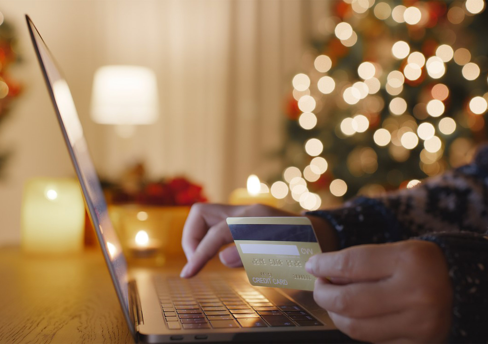 Person in a holiday sweater at their laptop with festive lights in the background using their Dover Federal Credit Union credit card to track and monitor holiday expenses.